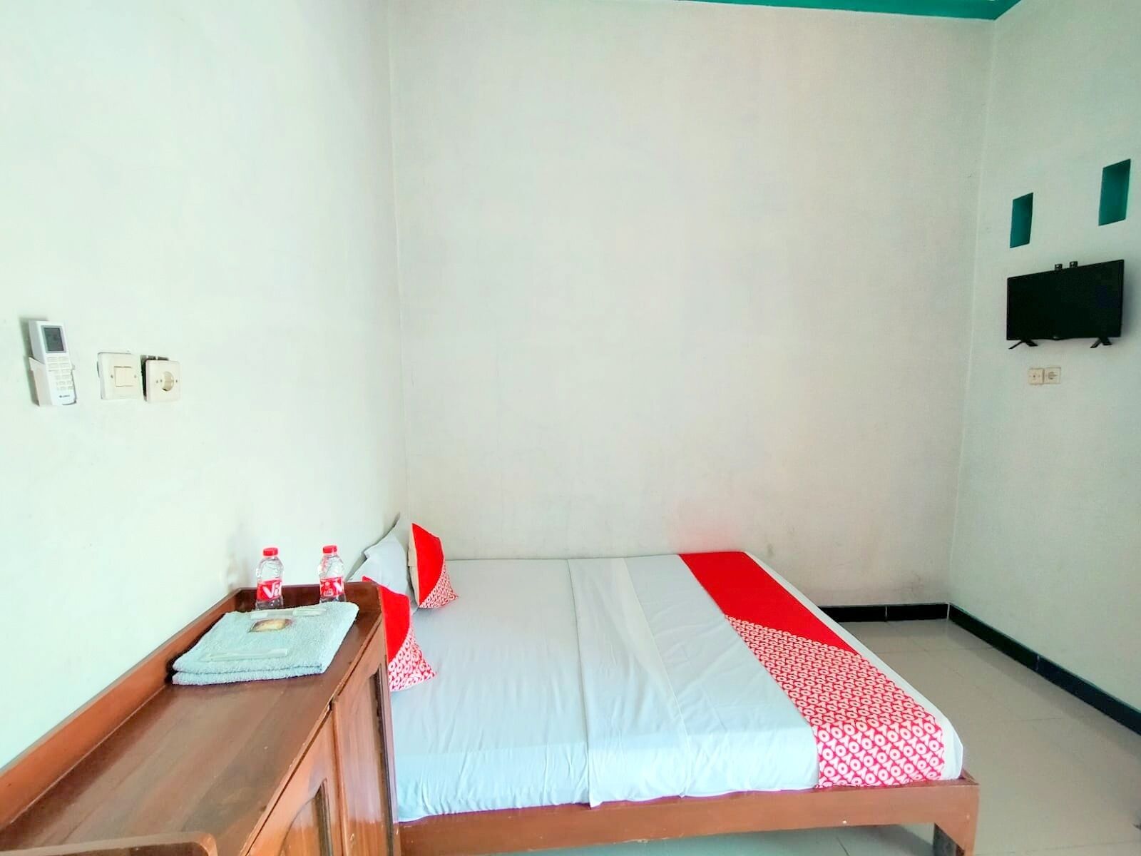 A small, simple room with a bed, a wooden nightstand, a TV mounted on the wall, and a few wall sockets.
