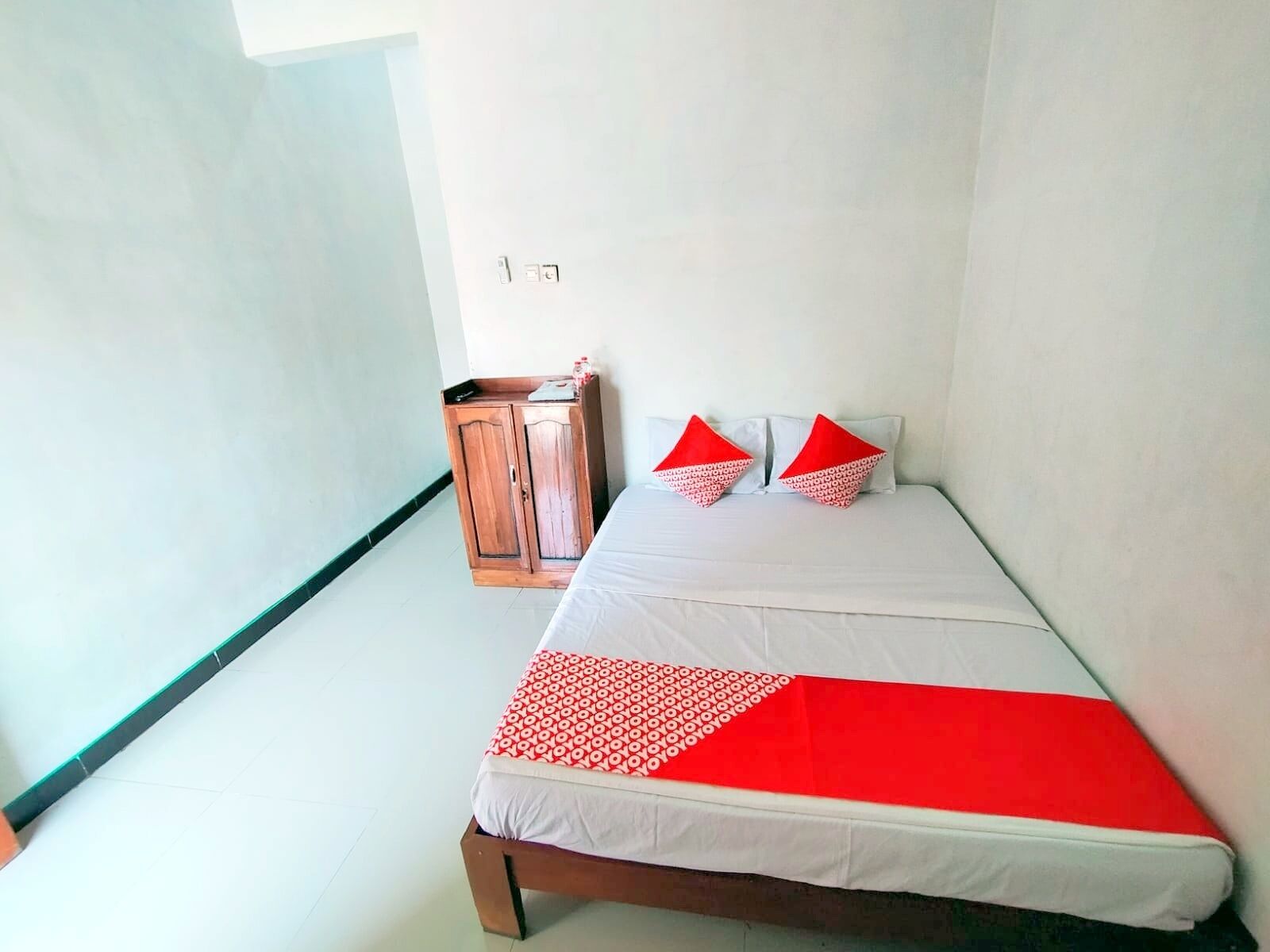 A single-bed room with a wooden wardrobe, a white bedspread, and two red pillows.