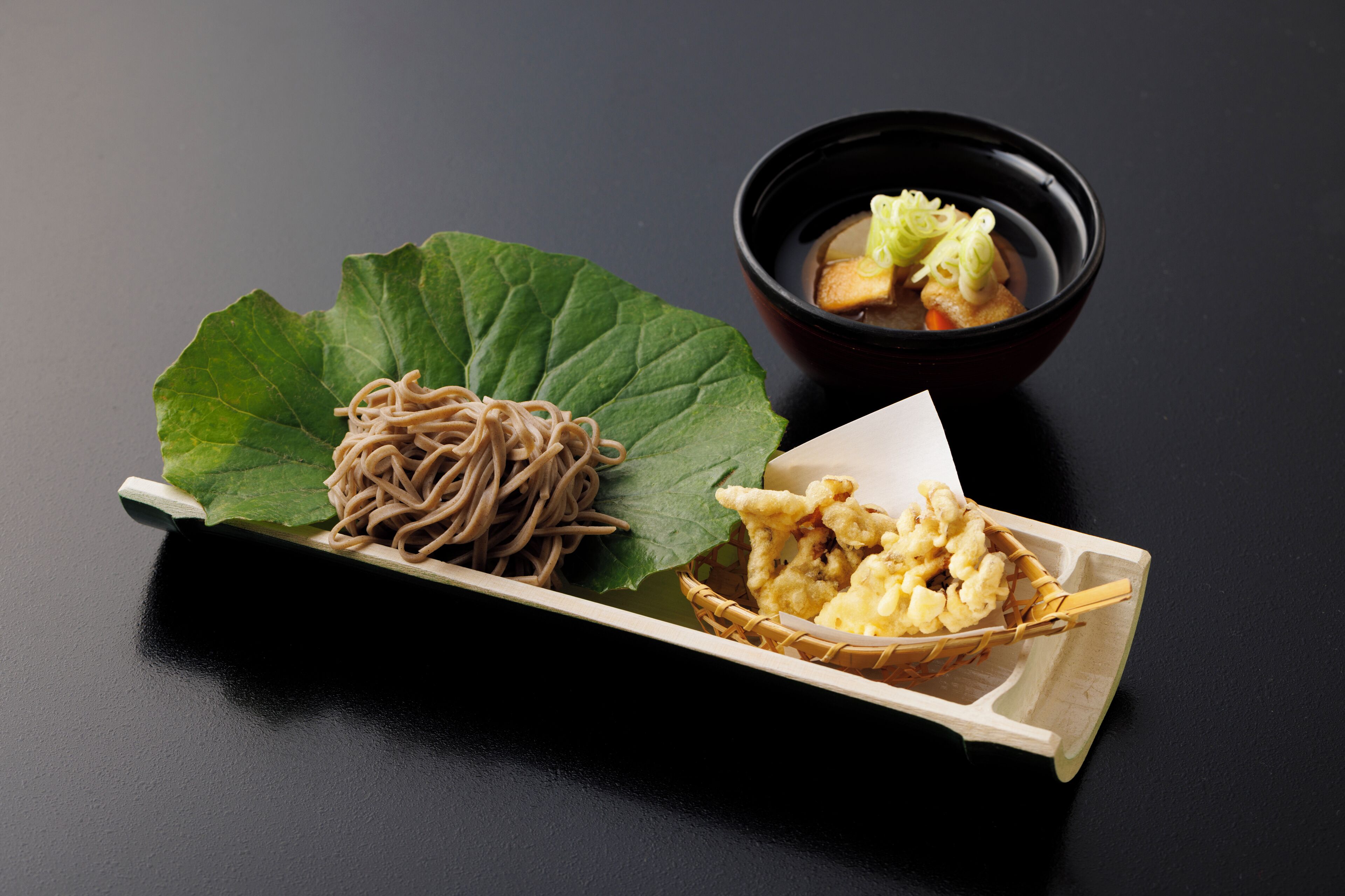 Un plato de fideos soba con una hoja verde, un tazón pequeño de sopa y una cesta de tempura sobre una superficie oscura.