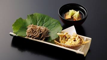 Un plato de fideos soba con una hoja verde, un tazón pequeño de sopa y una cesta de tempura sobre una superficie oscura.