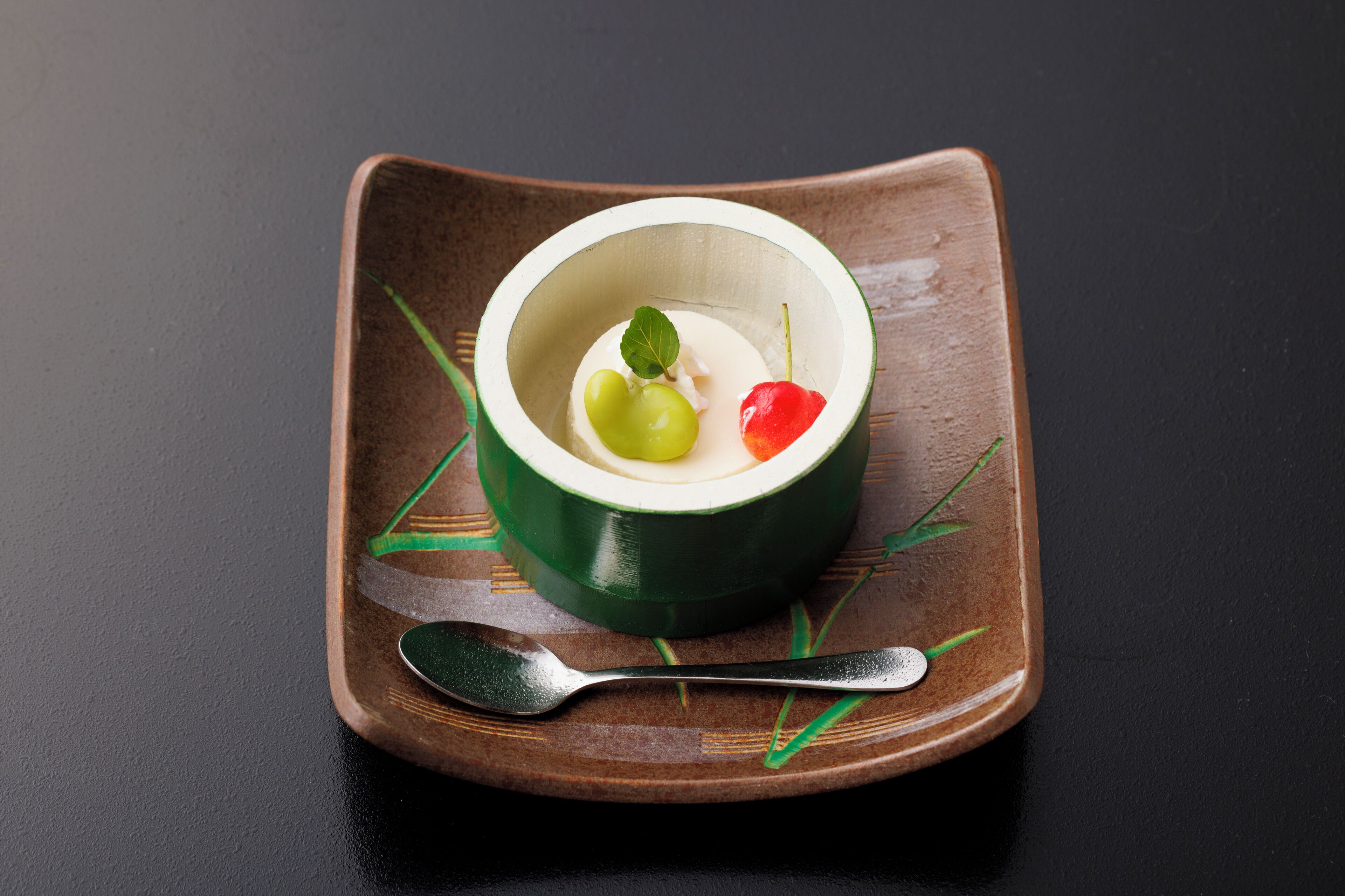 Un postre en un tazón verde sobre un plato cuadrado, con una cuchara al lado.