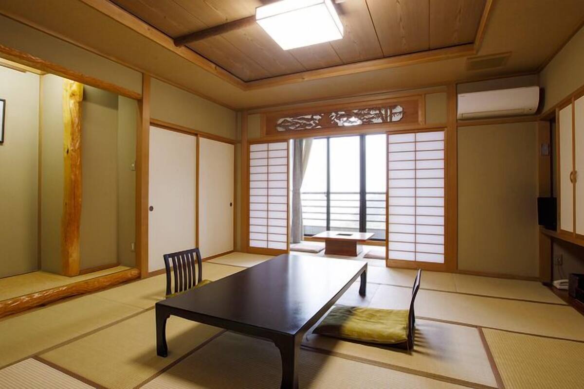 Una habitación japonesa tradicional con suelo de tatami, una mesa baja, una silla, puertas correderas y vistas a través de biombos shoji.