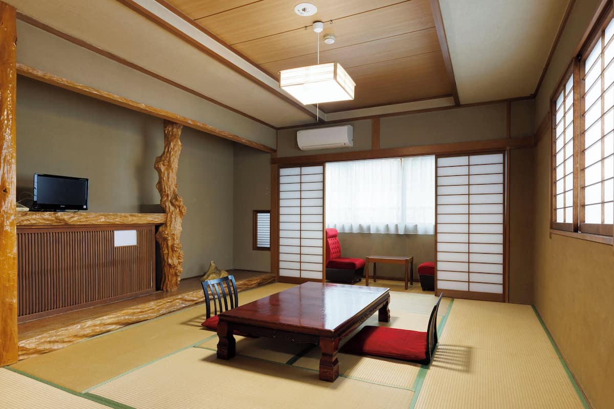 Una habitación japonesa tradicional con suelo de tatami, una mesa de madera baja, una silla roja y un televisor sobre un mueble de madera.