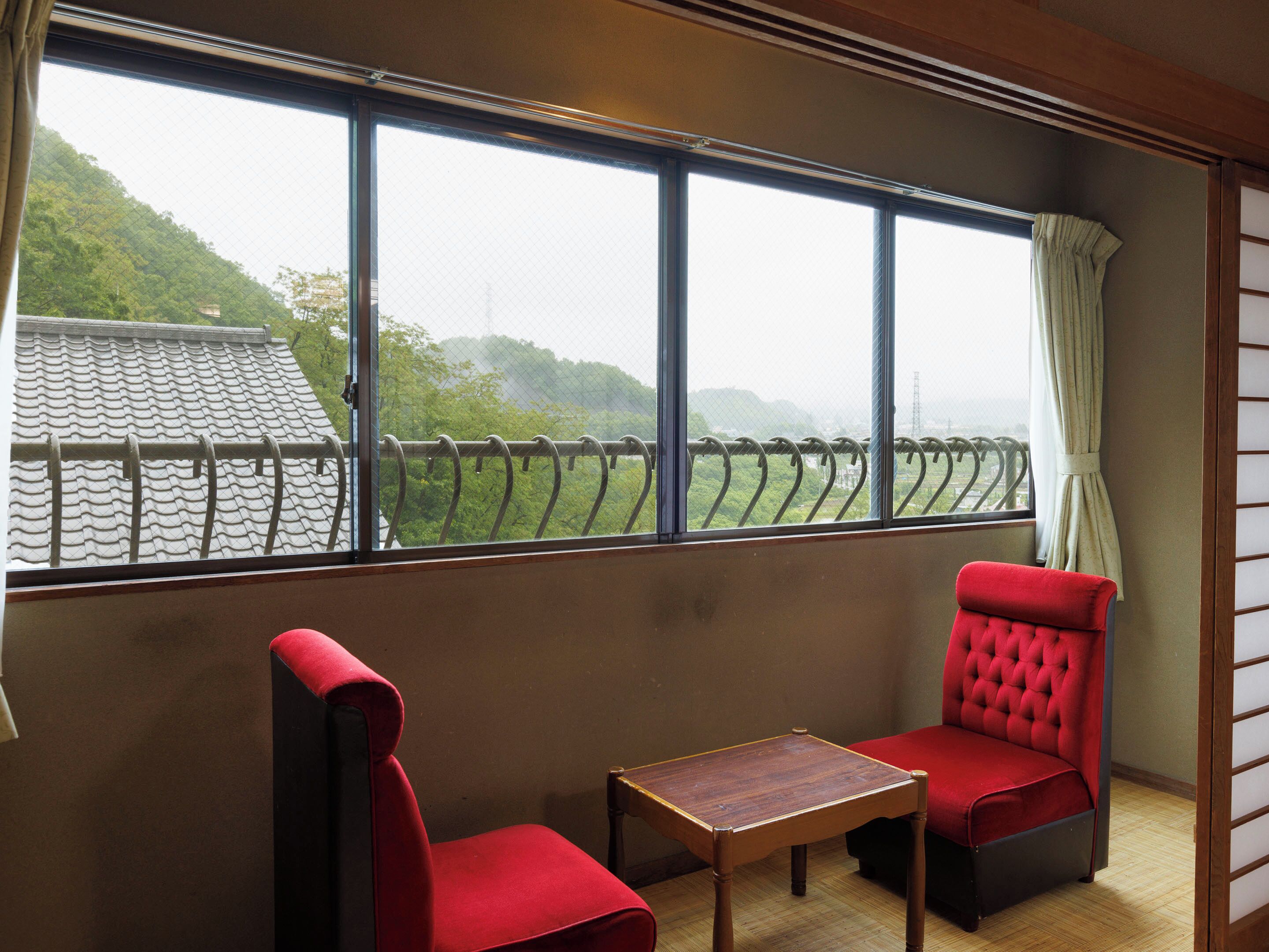 A room with a large window, two red upholstered chairs, and a small wooden table.