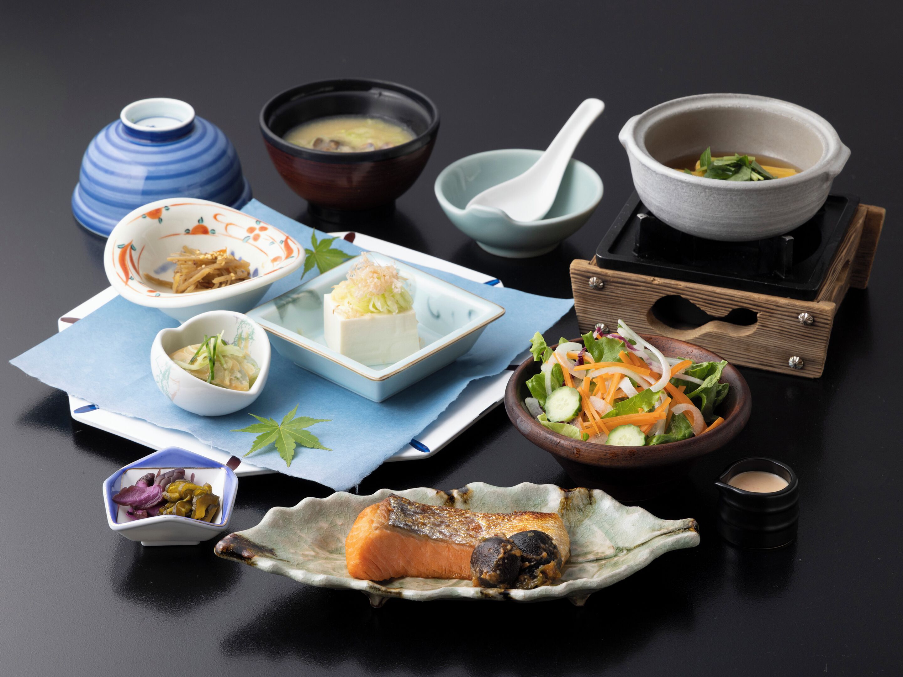 Forskellige retter, herunder en skål suppe, sashimi på en tallerken og en salat, anrettet på et bord med mørk baggrund.