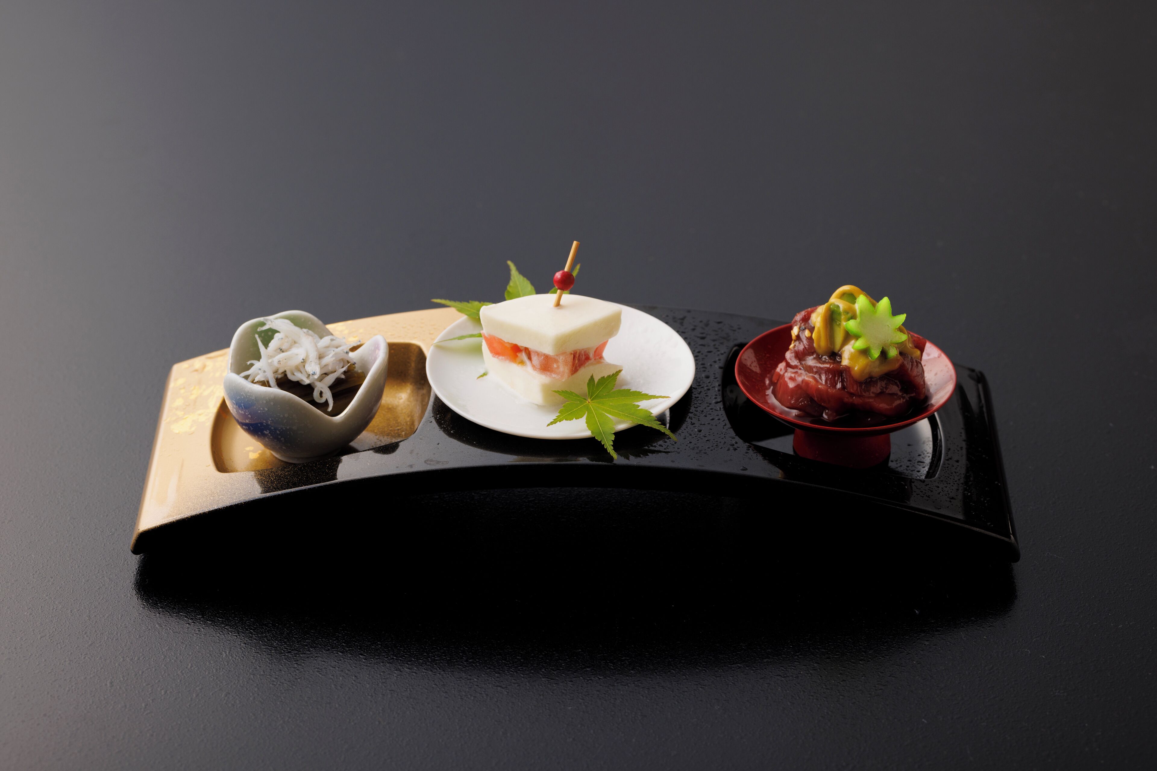 Una bandeja negra con tres platos: un tazón de sustancia blanca desmenuzada, un plato con un cubo blanco y una baya roja, y un plato rojo con guarnición verde.