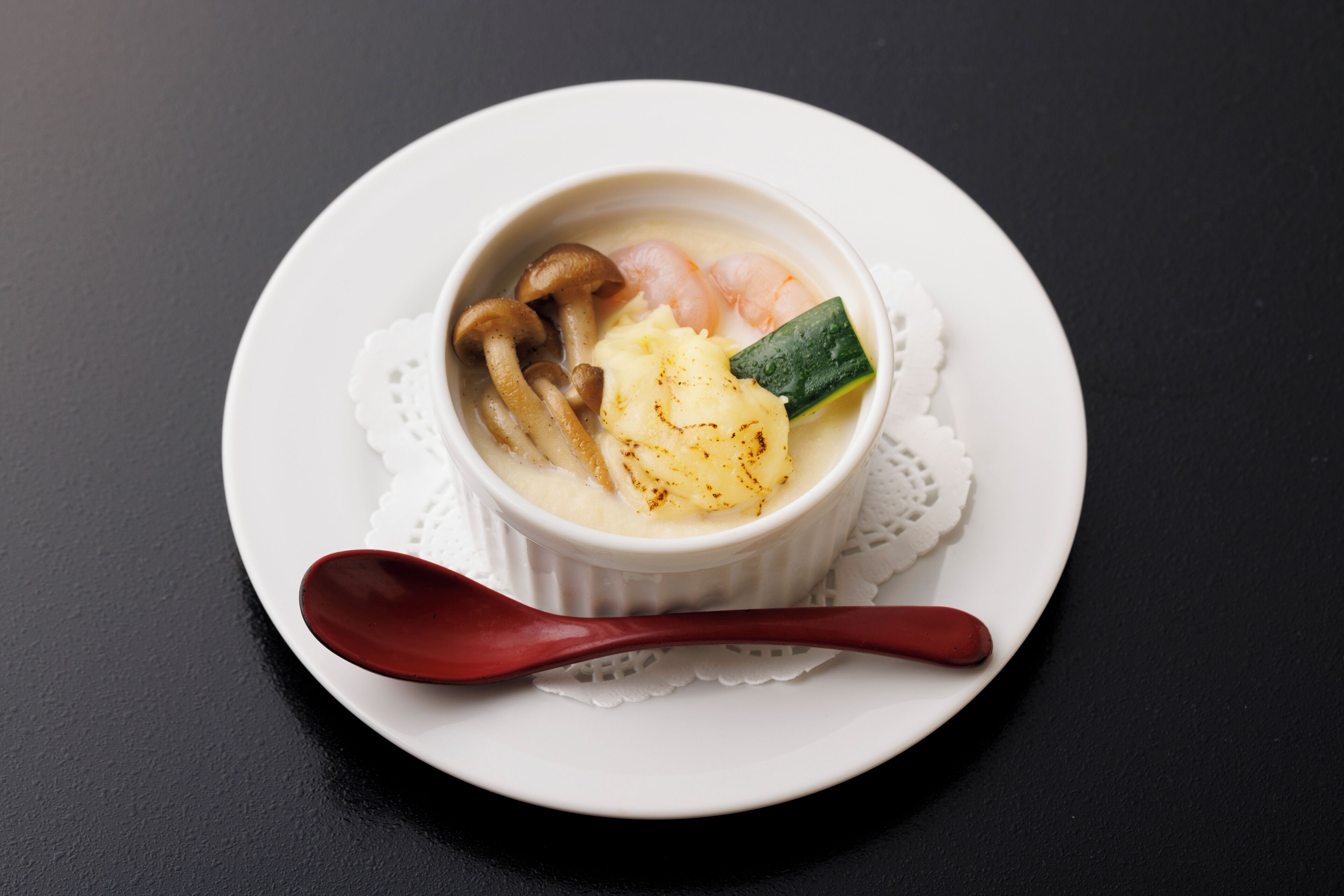 Un tazón de sopa con camarones, champiñones y calabacín sobre un plato blanco con una cuchara roja.