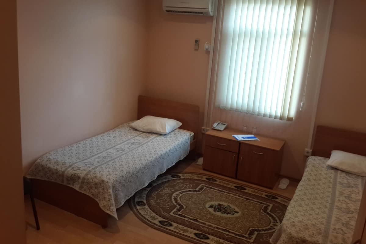 A small, single-bed room with a wooden headboard, a bedside table with a phone and glass, and a window with blinds.