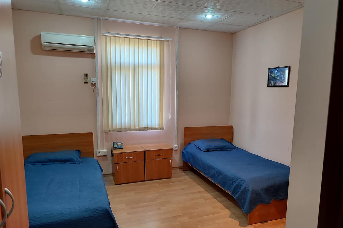 A small, clean room with two single beds, a wooden dresser, a window with blinds, and an air conditioning unit.