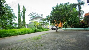 A paved pathway with a brick pattern, surrounded by greenery and a parked car. - OYO 2415 Ibma Smart Syariah (Pandaan)