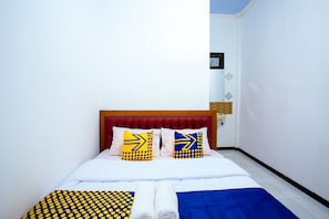 Uma cama bem arrumada com cabeceira vermelha, duas almofadas com padrões geométricos e um cobertor azul e amarelo com bolinhas.