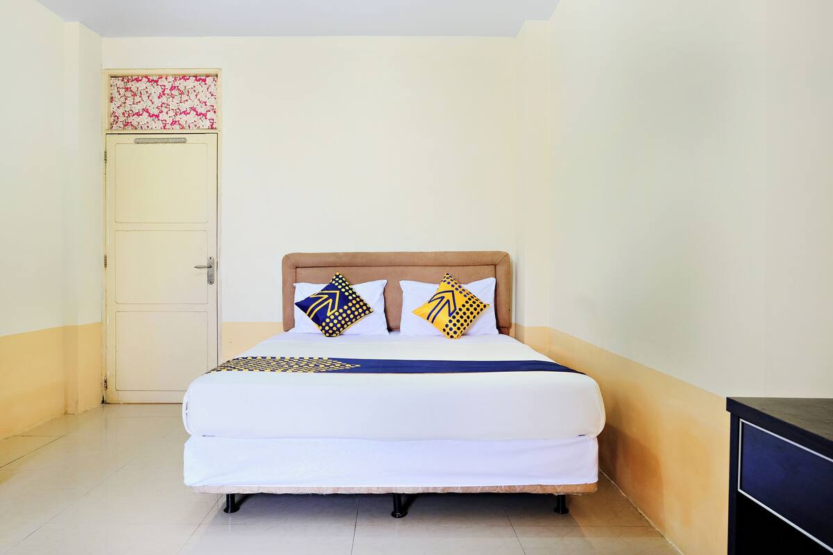 A single bed with a wooden headboard, two pillows with geometric patterns, a blue and white bedspread, and a plain door with a floral-patterned top.