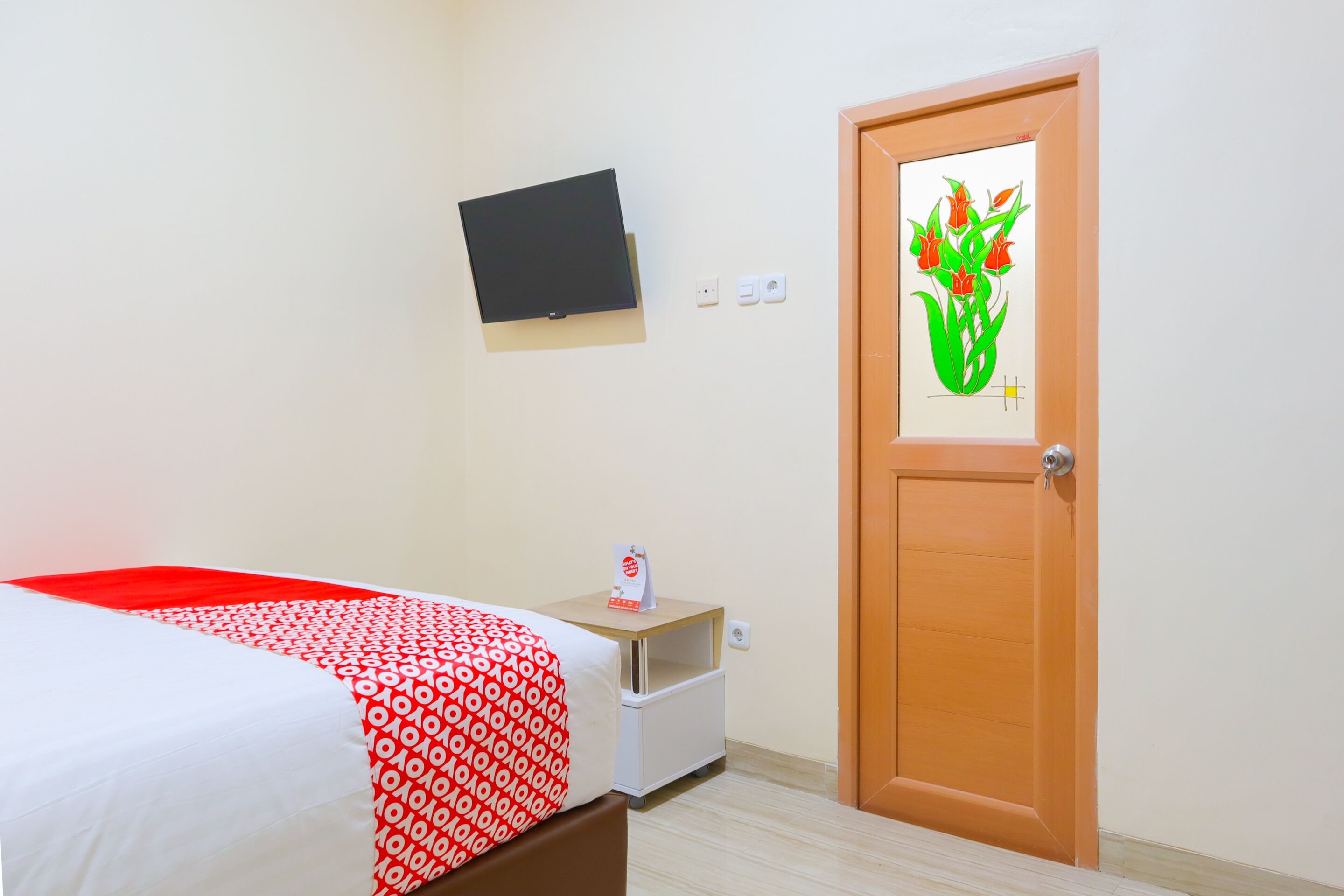 Una habitación de hotel con una cama, un televisor montado en la pared, una puerta de madera y una mesita de noche con un cartel.