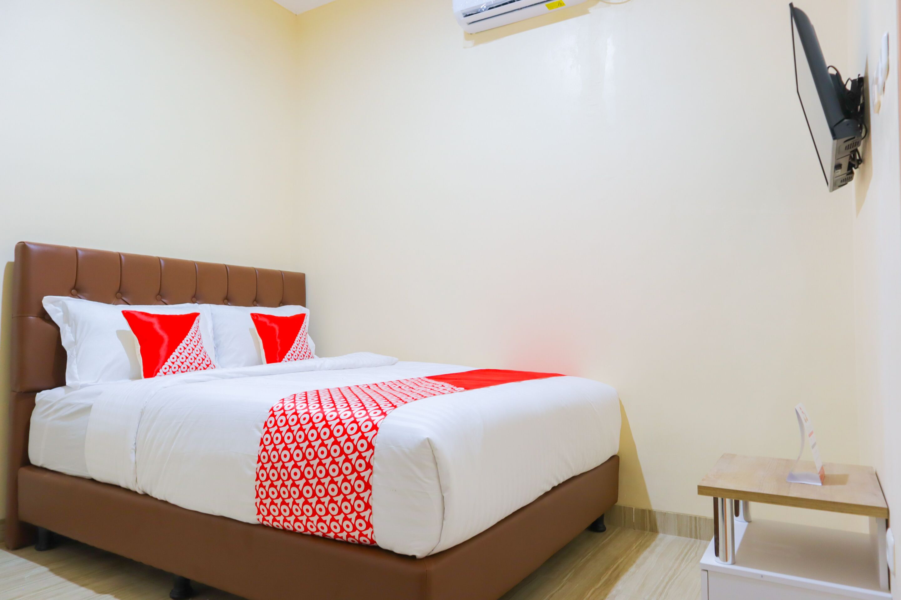A neatly made bed with a brown headboard, white and red bedding, and a small bedside table with a lamp.