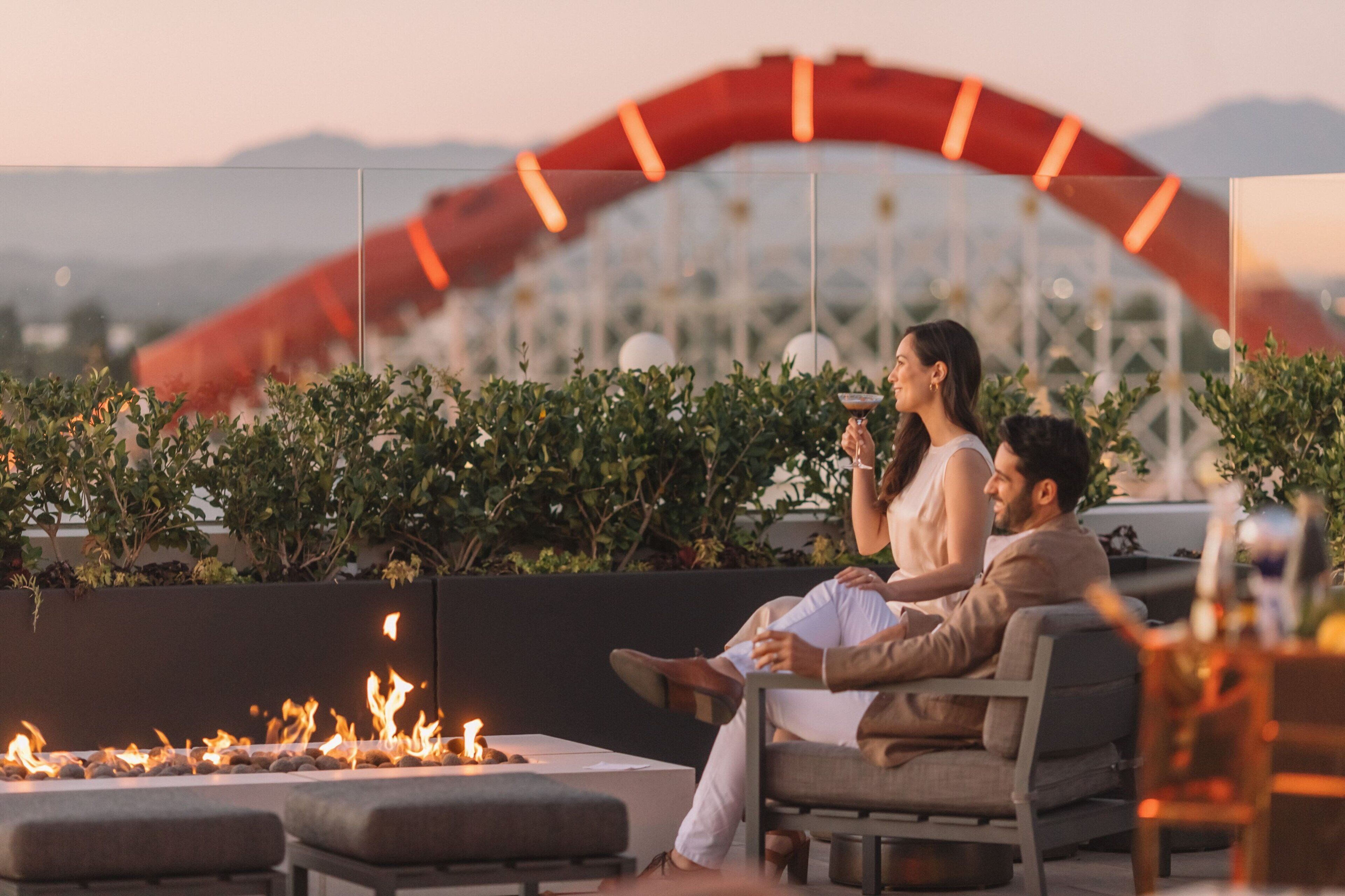A couple sitting by a fire pit in an outdoor seating area with a view of a cityscape and mountains in the background.