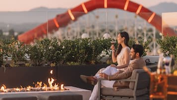 A couple sitting by a fire pit in an outdoor seating area with a view of a cityscape and mountains in the background.