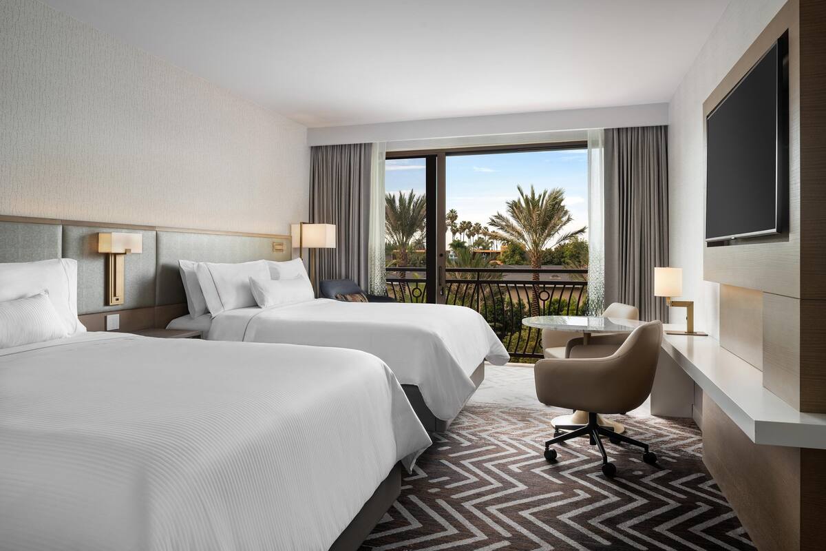 Habitación de hotel con dos camas, un escritorio, una silla y un balcón con vista a palmeras.