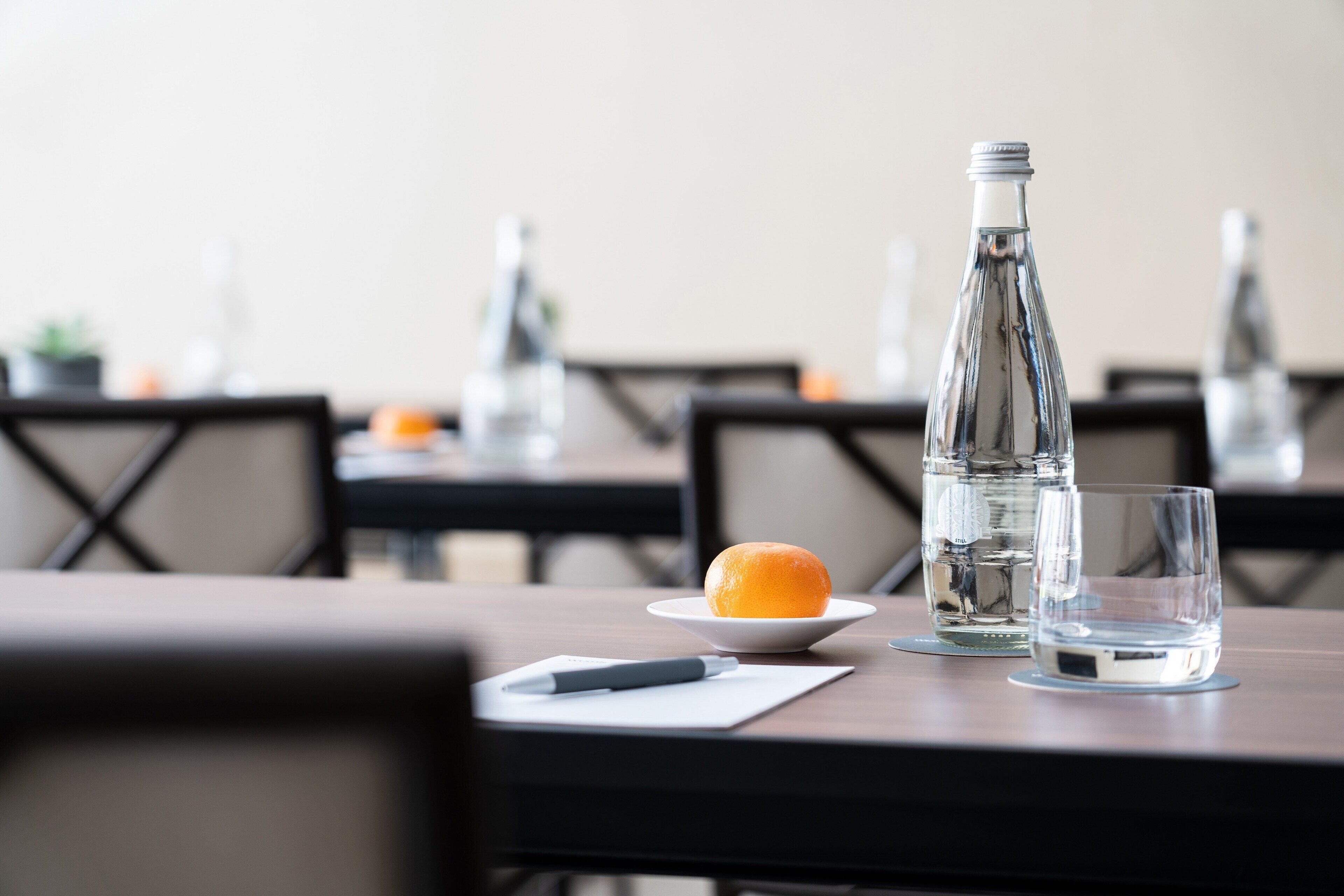 Una mesa de comedor con una botella de agua, un vaso, un plato con una naranja y un bolígrafo sobre una servilleta.