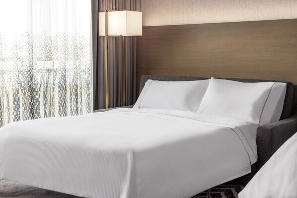 A neatly made bed with white linens, a bedside lamp, and a view of trees through sheer curtains. - The Westin Anaheim Resort (Anaheim)