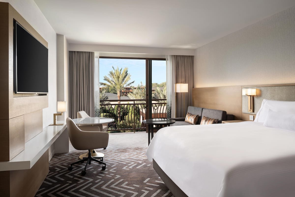 Habitación de hotel con una cama grande, un escritorio con silla, una mesita y un balcón con vista a palmeras y edificios.