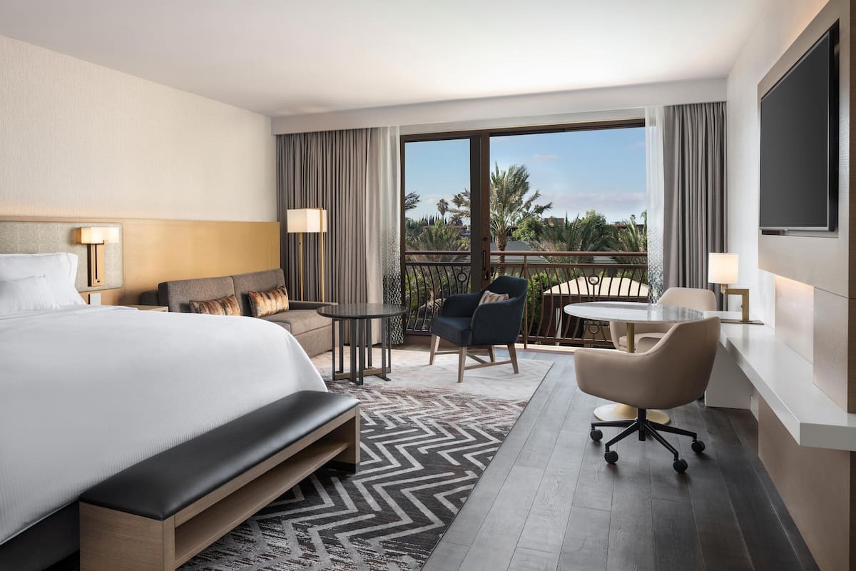 Habitación de hotel con una cama grande, un escritorio, una silla, una mesita y un balcón con vistas a palmeras.