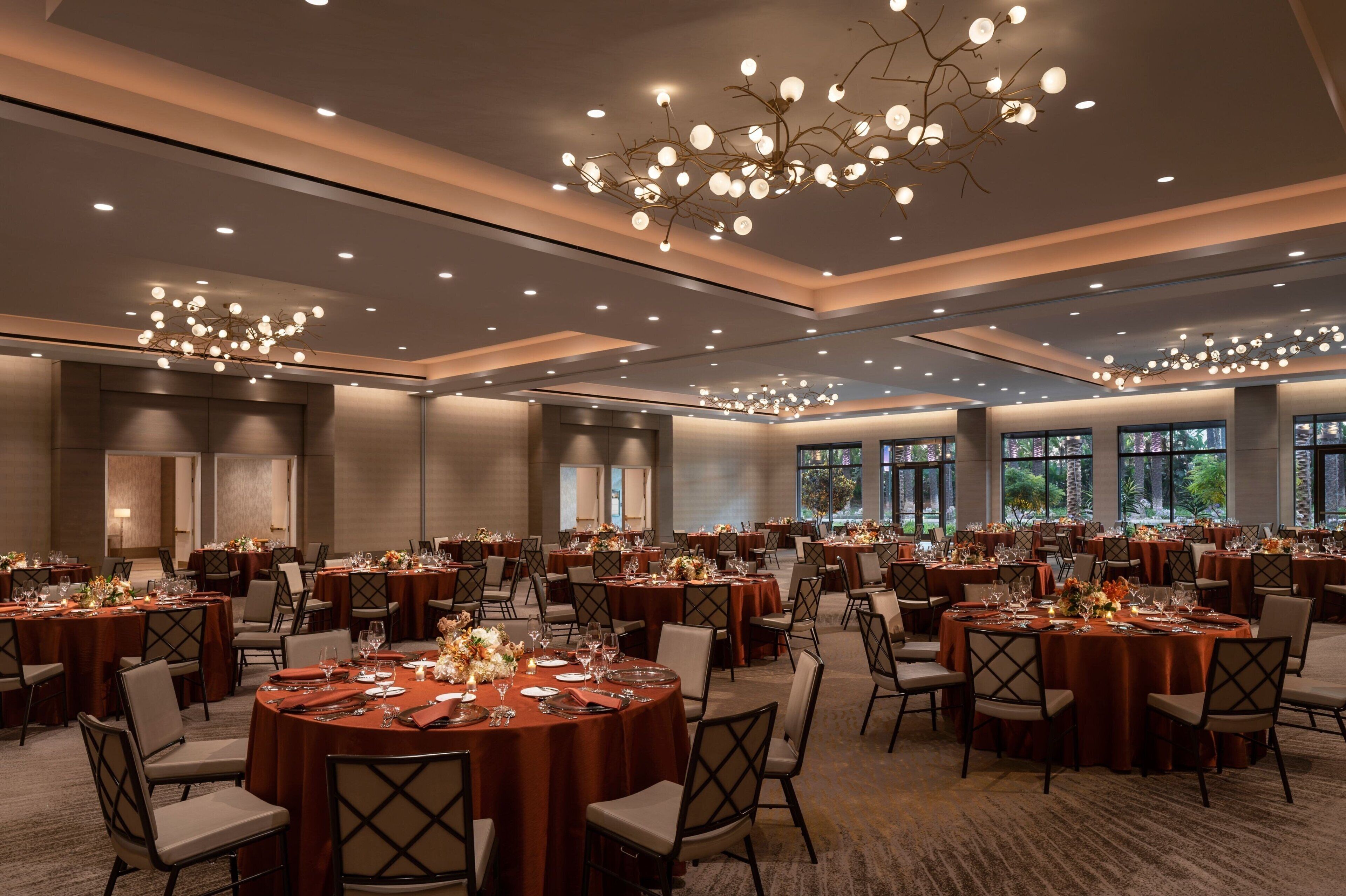 A large banquet hall with round tables set for dining, featuring overhead lighting and large windows.