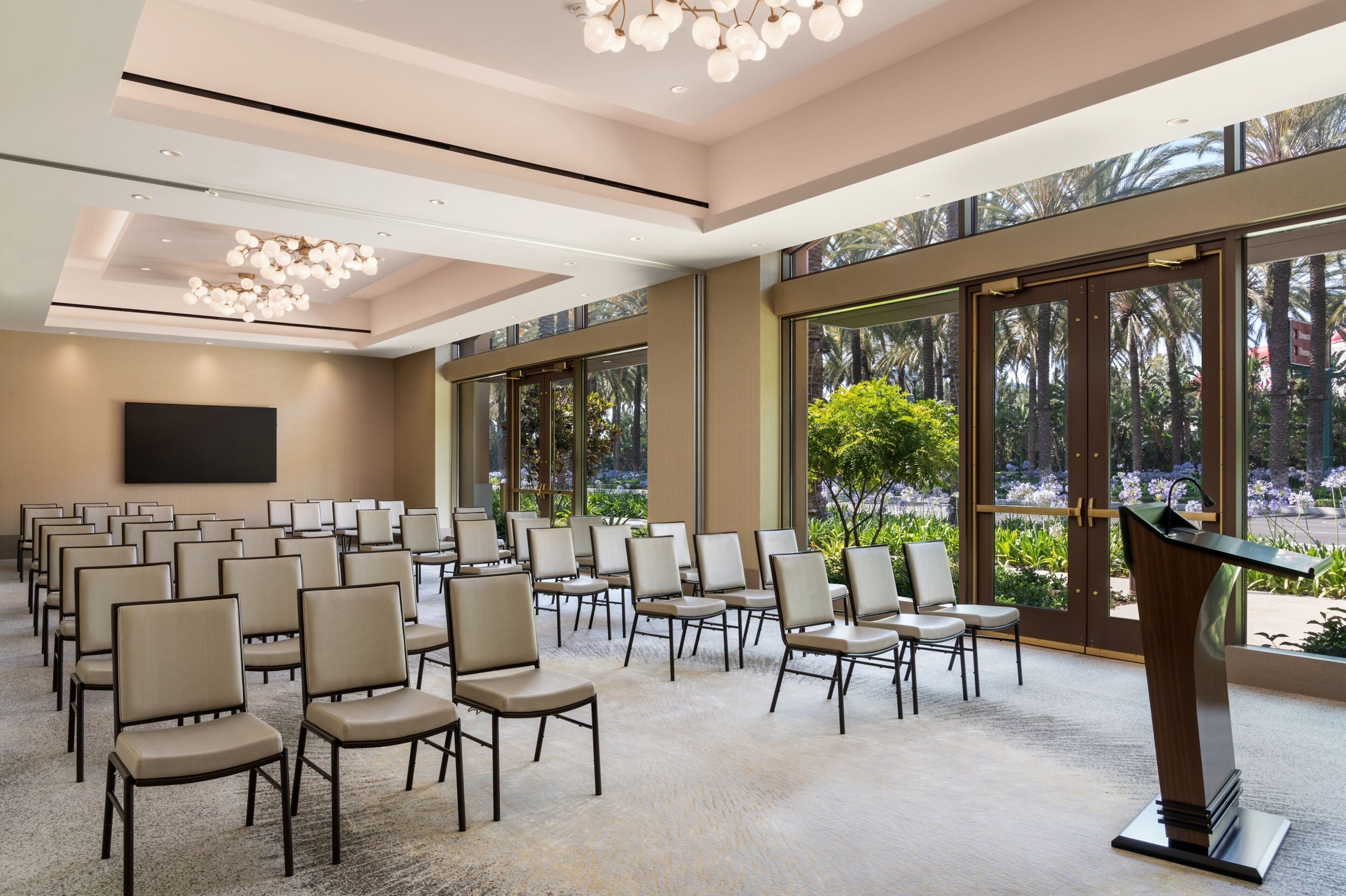 A conference room with rows of chairs, a podium, and large windows offering a view of palm trees.