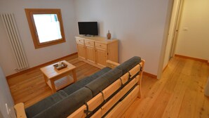 A small, cozy room with a wooden floor, a sofa, a TV on a wooden cabinet, and a window with blinds. - Beautiful apartment for 3 guests with WIFI, TV, patio and pets allowed (Raveo)