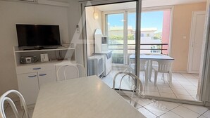 A kitchen with a TV, microwave, and toaster, a dining area with a table and chairs, and a sliding glass door leading to an outdoor patio with a table and chairs. - 1 CHAMBRE, DERNIER ETAGE AVEC ASC, CLIMATISATION (Sète)