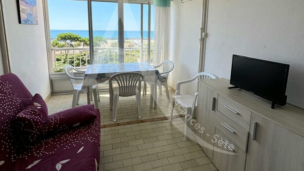 A room with a balcony, a dining table with chairs, a sofa, a TV on a cabinet, and a view of the sea. - STUDIO 4 PERS AVEC PKG BORD DE PLAGE VUE MER (Sète)