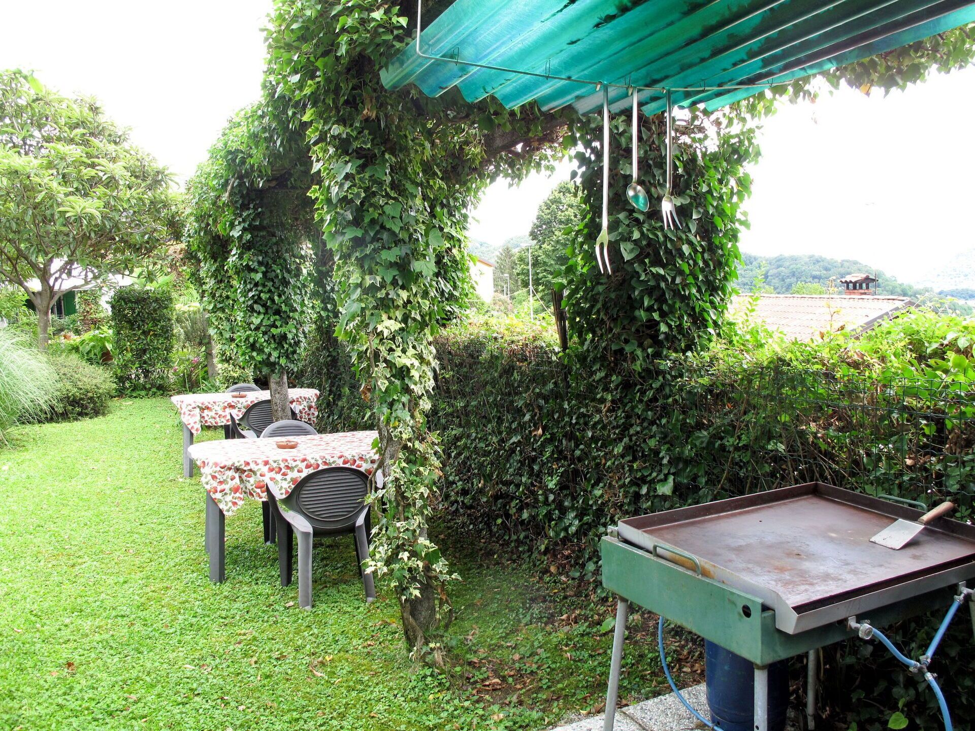 Un patio al aire libre con mesas cubiertas de manteles florales, rodeado de abundante follaje y una estación de parrilla de metal.