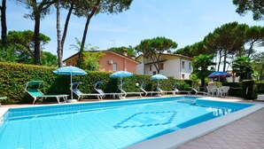 Una piscina con bordes de azulejos, rodeada de sillones de descanso y sombrillas, con un edificio al fondo.