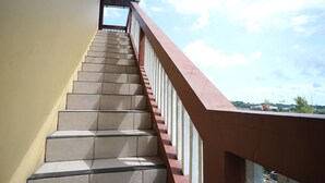 A staircase with beige steps and a red handrail, leading to a covered area with a view of the sky and distant buildings.