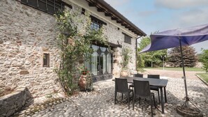 A stone building with a patio area featuring a table and chairs, a large umbrella, and a potted plant. - Cosy apartment in villa for 4 guests with A/C, pool, WIFI, TV and terrace (Povoletto)