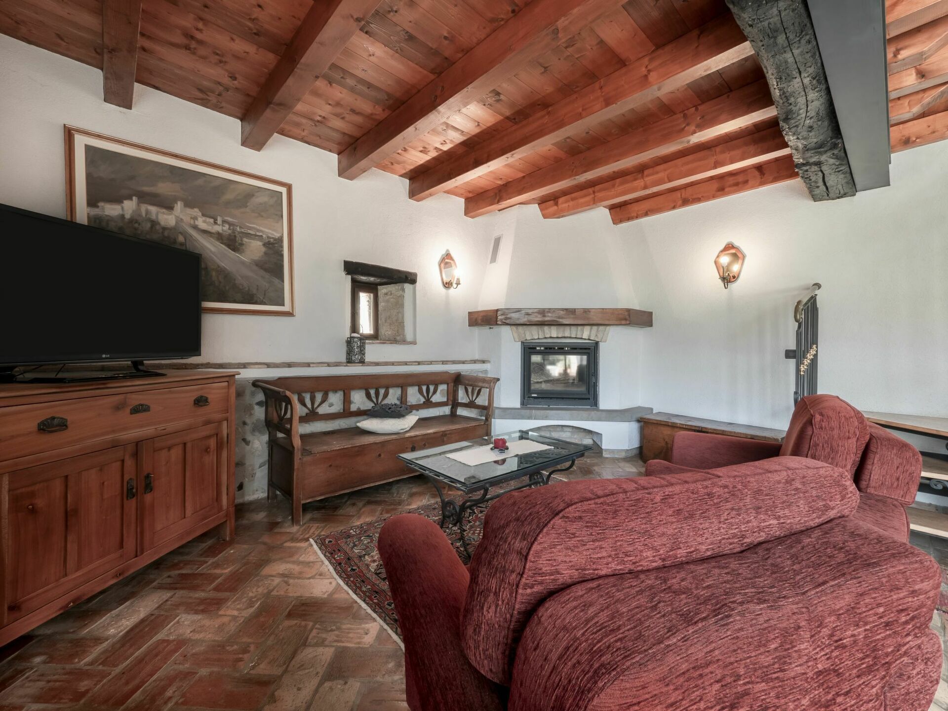 A cozy living room with a fireplace, wooden beams, a television, and a red sofa.
