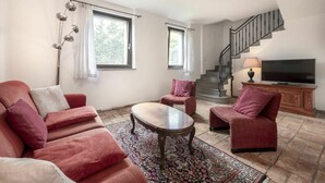 A living room with a sofa, armchairs, a coffee table, and a TV. There is a staircase with a railing and a lamp on a wooden cabinet. - Cosy apartment in villa for 4 people with A/C, WIFI, pool, TV, terrace and pets allowed (Povoletto)