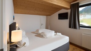 Ein Schlafzimmer mit Bett, Fernseher, Fenster mit Vorhängen, Holzboden und einer Tür.