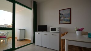 Eine kleine Küchenzeile mit einer Mikrowelle und einem Fernseher, ein Esstisch mit Stühlen, ein Blumentopf und ein Bild an der Wand.