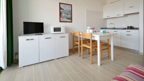 Eine kleine Küche mit weißen Schränken, einer Mikrowelle, einem Esstisch mit Stühlen, einem Fernseher auf einem Schrank und einem Bild an der Wand.