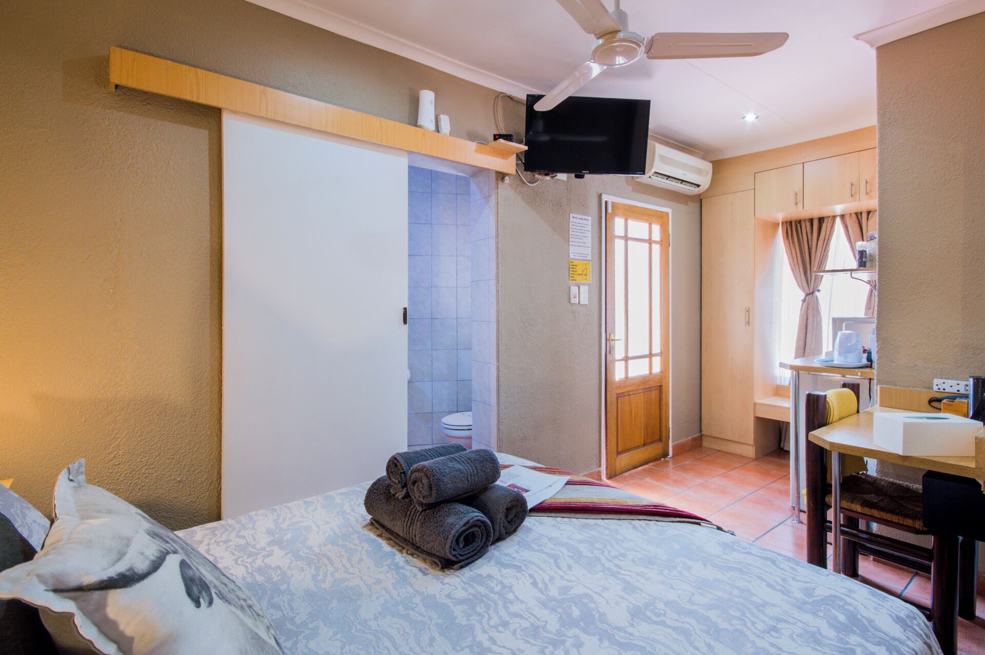 A hotel room with a bed, a desk, a chair, a ceiling fan, an air conditioning unit, and a television.