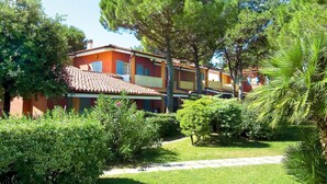A row of two-story buildings with a red-tiled roof, surrounded by lush greenery and a well-maintained lawn. - Nice apartment for 6 people with pool, A/C, TV, balcony and pets allowed (Bibione)