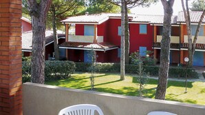 A balcony with white plastic chairs and a table, overlooking a courtyard with red buildings and trees. - Nice apartment for 6 people with pool, A/C, TV, balcony and pets allowed (Bibione)