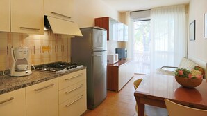 A compact kitchen with a gas stove, refrigerator, and microwave. There is a dining table with chairs and a small window with curtains. - Cosy apartment for 5 people with A/C, pool, TV, balcony and pets allowed (San Michele Al Tagliamento)