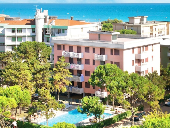 A hotel with a pool surrounded by trees, a view of the sea, and balconies with laundry hanging.