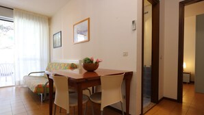 A dining area with a wooden table, four chairs, a bowl of flowers, and a window with curtains. - Cosy apartment for 5 people with A/C, pool, TV, balcony and pets allowed (San Michele Al Tagliamento)