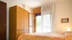 A bedroom with a wooden wardrobe, a bed with a colorful quilt, a window with curtains, and a bedside lamp. - Cosy apartment for 5 guests with A/C, TV, balcony and pets allowed (San Michele Al Tagliamento)