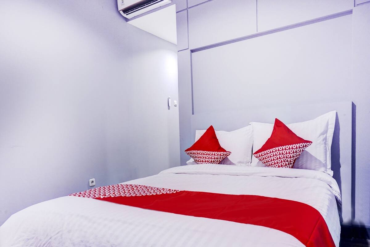 Une chambre d’hôtel moderne avec un lit soigneusement fait, des murs blancs et un plaid aux motifs rouges et blancs.