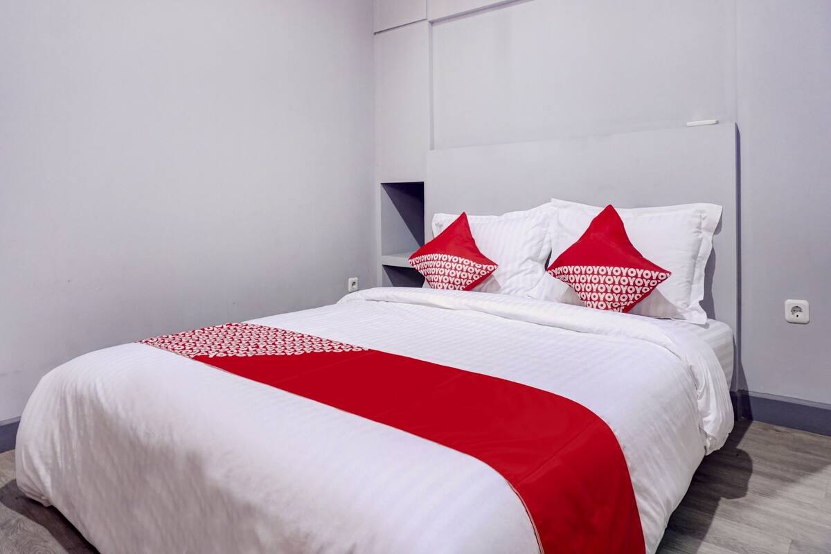Un lit bien fait, avec une literie blanche et rouge, une tête de lit dotée d’étagères intégrées et un mur blanc sobre.