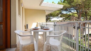 Un balcon avec une table et des chaises blanches, une petite plante en pot, et une vue sur des arbres et un bâtiment au loin.