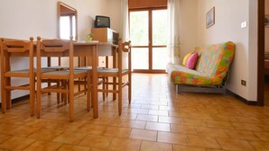 Une pièce comprenant une table et des chaises en bois pour la salle à manger, un canapé coloré, un téléviseur posé sur un meuble et une grande fenêtre avec des rideaux.